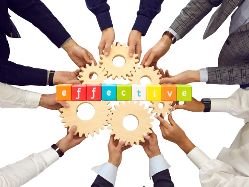 A group of diverse hands collaborates to hold interlocking wooden gears and colorful blocks spelling "effective" against a white background, symbolizing teamwork and efficiency—qualities essential for crafting SEO-friendly meta descriptions in small businesses.